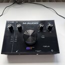 Černý audio rozhraní s nápisem M-AUDIO a označením modelu 192|4. Na předním panelu je velký centrální ovládací knoflík, čtyři menší knoflíky a logo a…