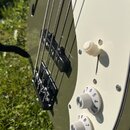 Detail elektrické basové kytary v olivově zelené barvě s krémovým pickguardem. Jsou vidět dva bílé otočné knofíky s nápisy VOL a TONE a jeden bílý př…
