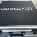 Černý transportní kufr s kovovými hranami a úchytem. Na horní straně je bílý nápis th.mann. Povrch vykazuje drobné nečistoty a opotřebení.
