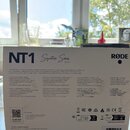 Fotografie znázorňuje kartónové balení mikrofonu. Na krabici je bílý nápis NT1 Signature Series BLACK a logo RØDE. Spodní část obalu obsahuje technic…