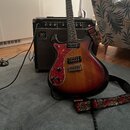 Elektrická kytara s trupem v červeno-žlutém sunburst provedení a červeným vzorovaným pickguardem. Na kytaru je nasazený černý popruh s červenými a bí…