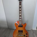 Elektrická kytara s trupem v žlutě-oranžovém sunburst provedení a hnědým hmatníkem s trapezoidními vložkami. Na hlavě je bílý nápis Gibson a kurzivní…