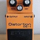 Oranžový kytarový efekt s nápisy Distortion a DS-1. Na horní části jsou tři ovládací knoflíky s popisky TONE, LEVEL a DIST a červená kontrolka CHECK.…