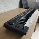 Černý digitální klavír s bílými a černými klávesami, umístěný na dřevěném stole. Na horní části korpusu je bílé logo Roland a nápisy OCT TRANSPOSE, V…