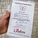 Fotografie zobrazuje papírový certifikát pravosti (Certificate of Authenticity) v průhledném plastovém obalu. Dokument obsahuje červené logo motýla a…