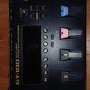 Černý kytarový procesor s nápisem BOSS a GT-100 AMP EFFECTS PROCESSOR. Zařízení obsahuje několik stoupacích spínačů s označením BANK UP, BANK DOWN, 1…