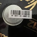 Detailní záběr na černý lesklý povrch s nápisy ve zlaté barvě, z nichž je čitelné „G12“ a část číslice „6“. Uprostřed je umístěn stříbrný kruhový prv…