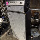 Černý basový kombozesilovač Ampeg umístěný na velké basové skříni. Horní jednotka má stříbrný ovládací panel s nápisy Ampeg, standby a power. Spodní…