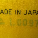 Detail dřevěného povrchu s nápisy. V horní části je černý tisk MADE IN JAPAN a pod ním zlatým písmem nápis SERIAL NUMBER L009791. Povrch vykazuje při…