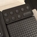 Černý ovládací panel s několika kulatými tlačítky a jedním oválným posuvníkem. Na tlačítkách jsou bílé symboly pro ovládání zvuku, přehrávání a další…