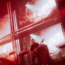 Fotografie zachycuje hudebníka na pódiu s basovou kytarou v tmavém provedení. V pozadí je vidět velká LED stěna s obrazem z koncertu a za hudebníkem…