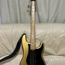 Zlatá elektrická basová kytara s černým pickguardem a javorovým hnedým hmatníkem. Na hlavě nástroje je nápis Fender Precision Bass. Kytara má čtyři s…