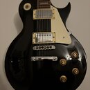 Černá elektrická kytara s tvarem single-cutaway a krčkem z tmavého dřeva. Nástroj má dva humbuckerové snímače s kovovými kryty, bílý plecháč a čtyři…