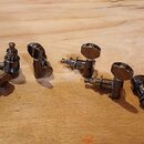 Šest kusů ladících mechanismů pro strunný nástroj s chromovaným povrchem. Jsou rozloženy na světlém dřevěném podkladu. Na mechanismu vlevo je viditel…