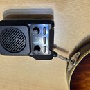 Černý kompaktní zesilovač s mřížkou reproduktoru, který je kabelem připojen k hnědé elektrické kytáře. Na horní straně jsou dvě otočné kontrolky s ná…
