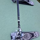 Dvojpedál na bicí soupravu s černými stupni a kovovým mechanismem. Na obou stupních je bílý nápis IRON COBRA 200 a logo TAMA. Povrch stupňů vykazuje…