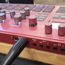 Červený sampler s nápisem electribe sampler MUSIC PRODUCTION STATION. Zařízení má několik ovládacích knoflíků, tlačítek a displej v pravé horní části…