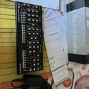Černý syntezátor s nápisem SE-02 STUDIO ELECTRONICS a logem Roland. Vedle přístroje jsou položeny bílé papírové doklady, černý napájecí adaptér a čás…