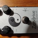 Stříbrný kytarový efekt s nápisem Zendrive a menším textem Hermida Audio USA. Na horní straně jsou čtyři černé otočné knoflíky s popisky Gain, Voice…