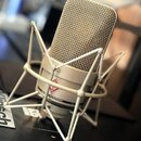 Kondenzátorový mikrofon béžové barvy s kovovou síťovkou, umístěný v odpruží stejné barvy. Na těle mikrofonu je viditelné logo s červeným symbolem a n…
