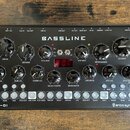 Černý syntezátor s nápisem BASSLINE a DB-01 v levém dolním rohu. V pravém dolním rohu je logo erica synths. Přístroj obsahuje několik černých otočnýc…