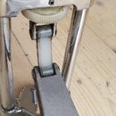 Detailní pohled na mechanismus pedálu z chromovaného kovu. Na horní části je vyražený nápis TIGHTEN. Viditelný je bílý plastový řemínek a šedá kovová…