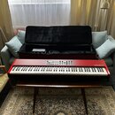Červený digitální klavír s bílými a černými klávesami. Na pravé straně korpusu je bílý nápis nord piano. Nástroj leží na hnědém dřevěném stole před o…