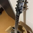 Černá elektrická kytara s logem Ibanez na hlavě. Na hlavě je rovněž bílý nápis Artist v kurzívě. Nástroj má hnědý hриф s bílými značkami na pražcích.…