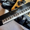 Černý klaviatura s bílými a černými klávesy. Na levé straně je nápis KOMPLETE s logem. V horní části se nachází displej, ovládací knoflíky a svislá t…