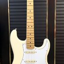 Krémově bílá elektrická kytara s jasným javorovým hnedlem krku a hmatníkem. Na hlavě je viditelné logo Fender. Kytara má tři snímače, bílý pickguard…