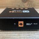 Černý kovový zdroj napájení s hranatým tvarem. Na zadní straně je umístěn vstup pro napájecí kabel a vedle něj nápis INPUT, 9V ~ 18V DC 2000mA. Na ho…