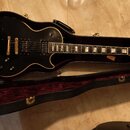 Černá elektrická kytara s tvarem singlecut v otevřeném kufru s purpurovým vnitřkem. Na hlavě je viditelné logo Gibson. Kytara má zlatý hardware a bíl…