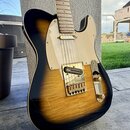 Elektrická kytara s tvarem typu Telecaster v provedení sunburst (černá okraje, žlutý střed). Nástroj má světlý hриф z javoru a krémový pickguard. Na…