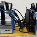 Dvě modrostříbrné studiové lampy značky ARRI s černými madly a klapkami. Na spodním bílém štítku obou přístrojů je uveden model T1 a logo ARRI. Povrc…