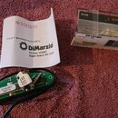Fotografie zobrazuje elektrický snímač s černým kabelem, plastové průhledné pouzdro a papírový vložek. Na snímači je vidět zelená deska s nápisem DP1…