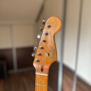 Fotografie zachycuje hlavu elektrické kytary z přírodního dřeva s šestimi ladicími kolíky. Na hlavě je vidět nápis Squier by Fender a STRATOCASTER®.…