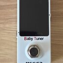 Bílý kytarový ladič v kompaktním obdélníkovém tvaru s černým displejem. Na přední straně jsou červeným nápisem „Baby Tuner“ a černým logem „MOOER“. V…