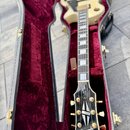 Bílá elektrická kytara v otevřeném černém kufru s červeným plyšovým vnitřkem. Na hlavě kytary je bílý ornament a nápisy „Gibson“ a „Les Paul Custom“.…