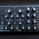 Černý semi-modulární analogový syntetizátor s dřevěnými bočními panely. Na spodní liště je bílý nápis SUBHARMONICON SEMI-MODULAR ANALOG POLYRHYTHMIC…