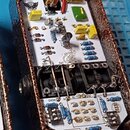 Fotografie zachycuje vnitřek efekt pedálu v hnědém kovovém šasi. Na bílé desce jsou rozmístěny elektronické součástky, včetně kondenzátorů s nápisy 3…