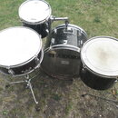 Sada černých bubnů z bicího setu umístěná na trávníku. Na předním obruči největšího bubnu je nápis ASAMA a pod ním menší text Professional Drum Equip…