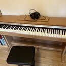 Digitální piano v dřevěném korpusu s černou klaviaturou a přiloženou černou stolečkou. Na horní desce nástroje leží černá sluchátka. Nad klávesami je…