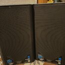 Dvě černé reproduktorové bedny s kovovou mřížkou. Na spodní části jsou viditelné nápisy K12.2, 2000 WATT a logo QSC s označením K.2 SERIES. Dále je z…