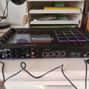 Černý MPC sampler s displejem a mřížkou barevně podsvícených padů. Na přední straně je nápis AKAI PROFESSIONAL a různé konektory s popisky jako MASTE…