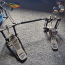 Dvojpedál na bicí buben s kovým provedením a černou spojovací tyčí. Na obou pedálech je viditelné logo MAPEX v oválném rámečku. Povrch pedálů vykazuj…