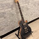 Elektrická kytara s tmavým, žilovatým povrchem těla a světle hnědými hranami. Na hlavě je viditelné logo značky Cort. Nástroj má dva humbuckery, tři…
