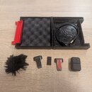 Sada příslušenství k mikrofonu v černém plastovém kufru s červeným prvkem a pěnovou výplní. Obsahuje černý USB kabel, malý černý bezdrátový vysílač s…
