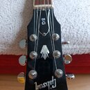 Černá hlava kytary s nápisem Gibson a označením SG. Na povrchu je vidět stříbrný ornament ve tvaru šipky a šest ladících kolíků. Povrch vykazuje drob…