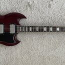 Tmavě červená elektrická kytara s černým pickguardem a dvěma humbuckery. Na hlavě je zlaté logo Vintage a dekorativní ornament. Nástroj vykazuje mini…