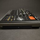 Černý digitální mixážní pult s kovovým stříbrným rámem. Na pravé straně je oranžový displej a nápis "TASCAM DP-24". Zařízení obsahuje řadu posuvníků,…